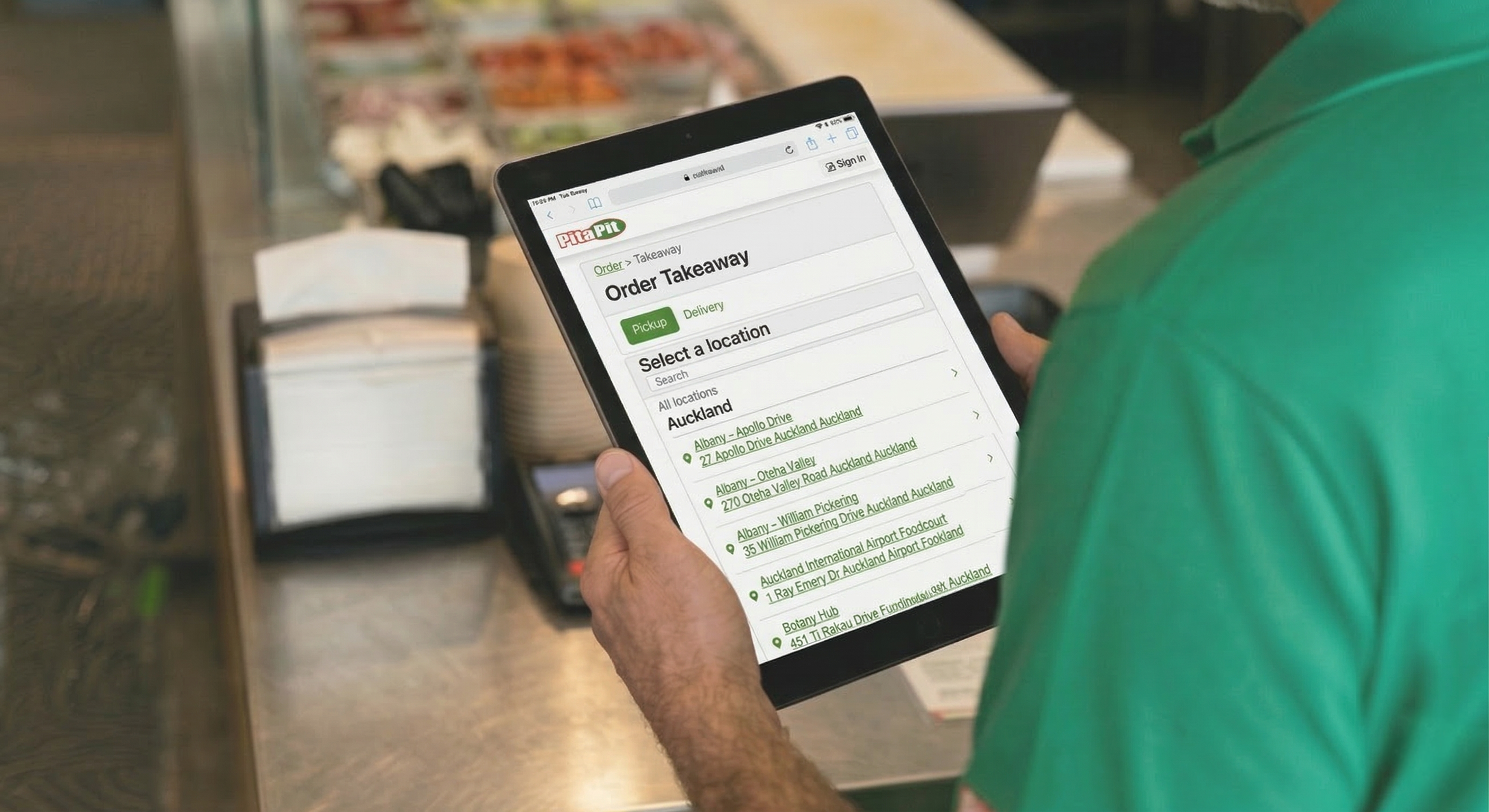 Restaurant staff member holding a tablet showing a multi-location ordering page for selecting a store.