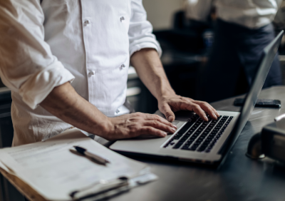 Restaurant staff reviewing sales reports on a laptop in a kitchen workspace.