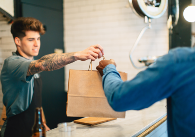 A tattooed staff member handing a takeaway bag to a customer across a cafe counter.