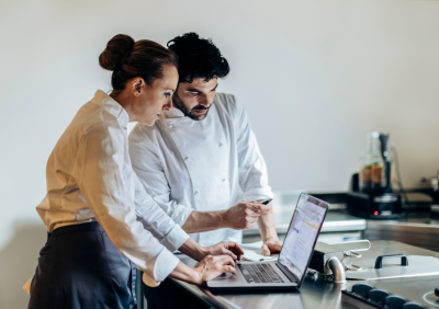 Restaurant staff reviewing sales reports on a laptop in a kitchen workspace.