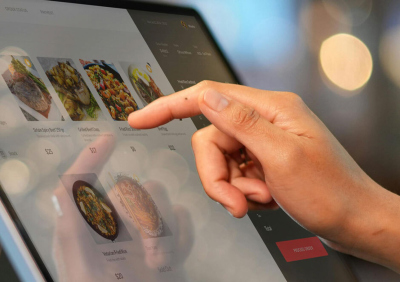 Close-up of a customer's finger selecting a menu item on a dark touchscreen food ordering kiosk.