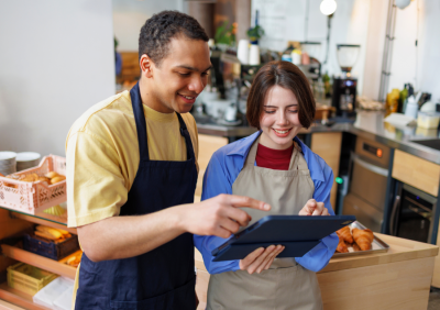 Cafe owner reviewing order analytics on a tablet behind the counter in a busy bakery cafe.